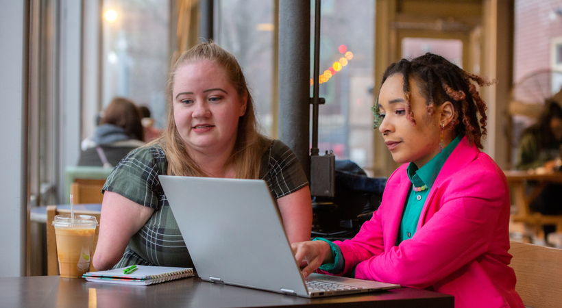 Two women are sat at a desk looking at a laptop.