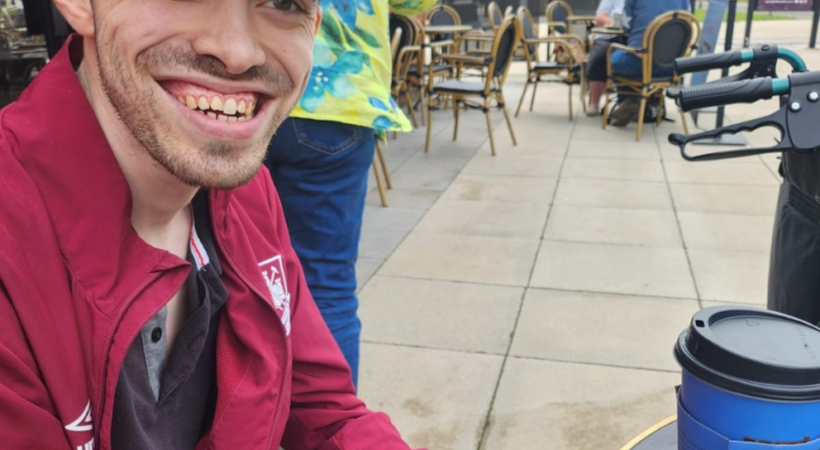 James is sat outside drinking a coffee. He is smiling at the camera.